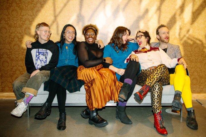 Six writers sit on a bench against a gold background facing the camera with arms around each other's shoulders laughing.