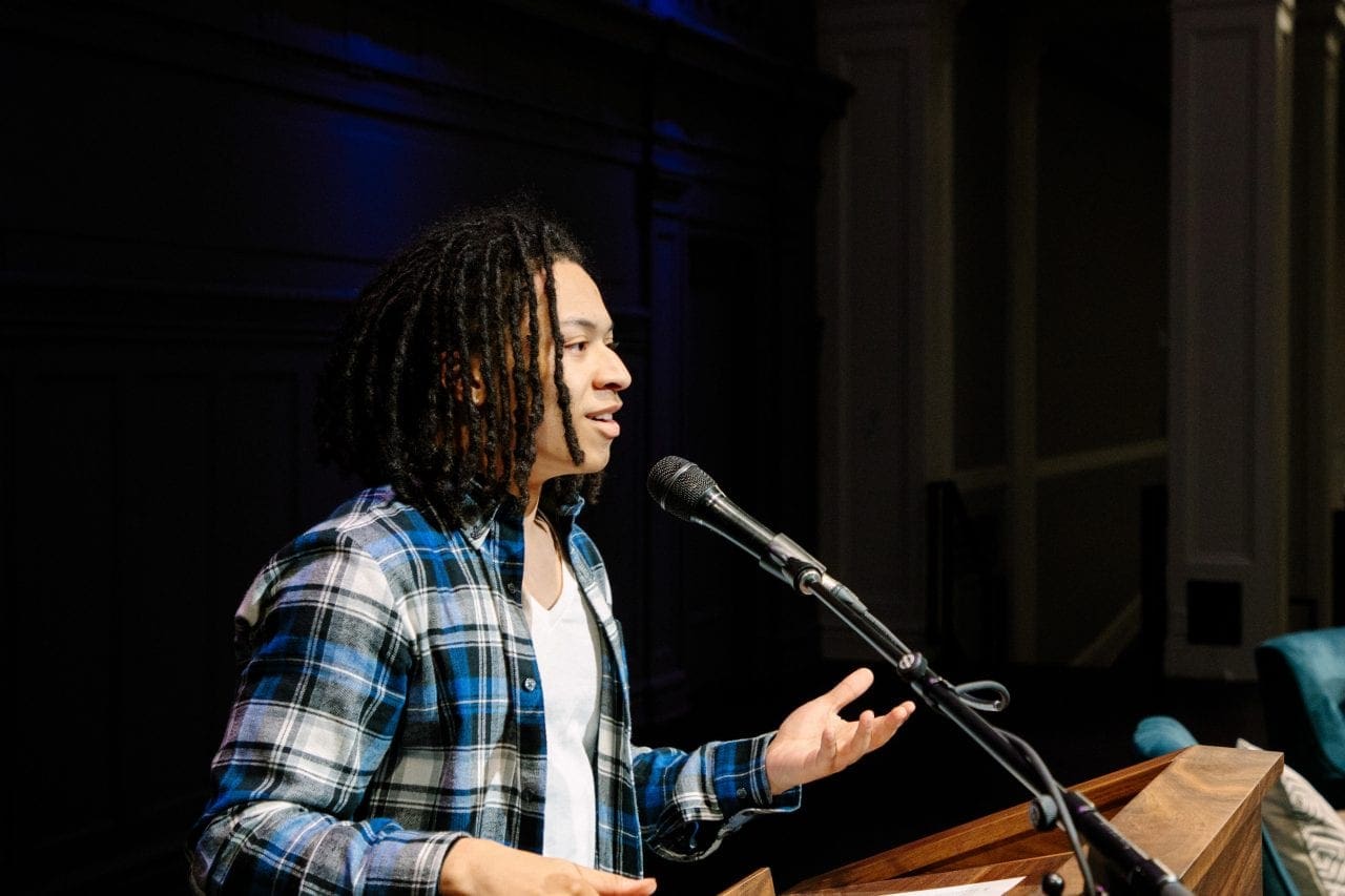 Gianni Johnson stands at the podium, smiling and speaking into a microphone, with one hand raised.