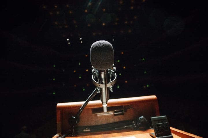 A microphone, standing atop a lectern, is lit by the glow of stage lights