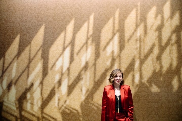 Paisley Rekdal, in a red satin suit, stands against a wall at Hugo House that is designed to look like the shadows cast by Emily Dickinson's bedroom window.