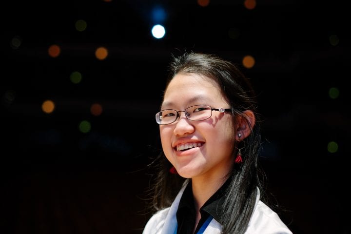 Wei-Wei Lee, looking slightly over her shoulder, grins at the camera amid a black backdrop lit by multicolored, circular lights.