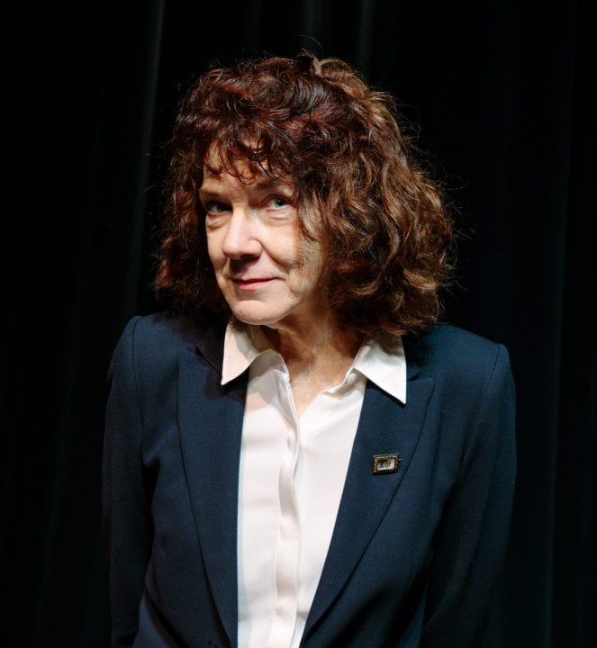 Mary Ruefle gazes into the camera, head down, brows up, through her curly hair. Her navy blue blazer is decorated with a small pin.