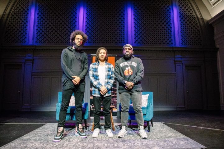 From left of right, Quenton Baker, Gianni Johnson, and Hanif Abdurraqib, stand on a dramatically purple-lit stage at Town Hall, with their arms all posed the same, clasped in front of their torsos. They all have serious expressions.