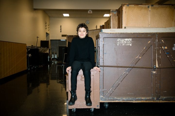A kid with curly hair sits atop a box backstage at Benaroya Hall, legs swinging.