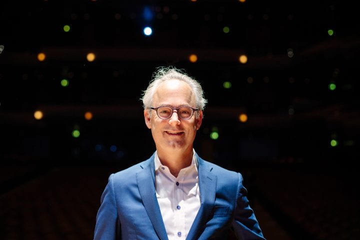 Amor Towles, in glasses and a bold blue suit, stands in the center of the picture, against a backdrop lit by multicolored, circular lights. He's smiling energetically and making direct eye contact.