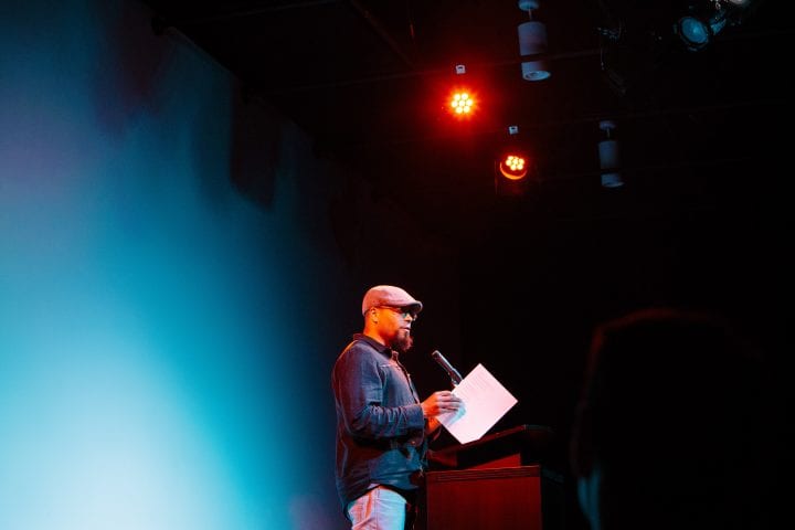 A man wearing a golf cap speaks into a microphone at a lectern. The stage lights cast a blue, artsy lens flare across half of the image.