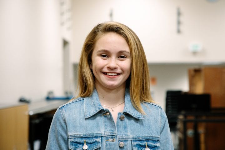 A young girl smiles into a camera backstage, wearing a silver necklace and denim jacket.
