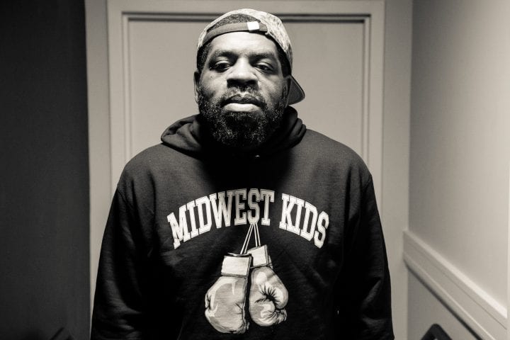 A black and white shot of a bearded Hanif Abdurraqib, framed in a door frame. He's wearing a graphic sweater with boxing gloves that says "Midwest Kids."