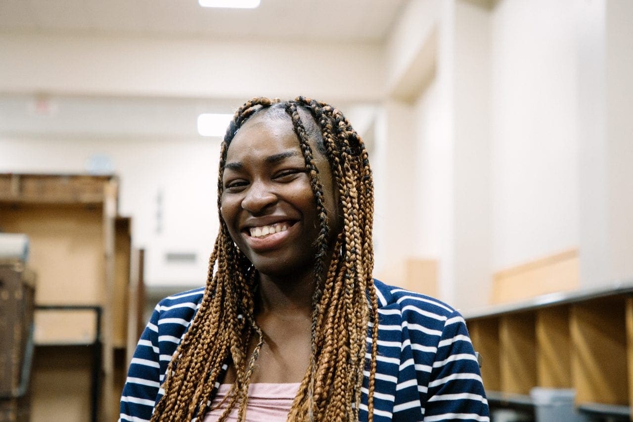 Chimamanda Danita Egboh laughs backstage at Benaroya Hall. Her hair is in braids, and she's wearing a blue and white striped cardigan.