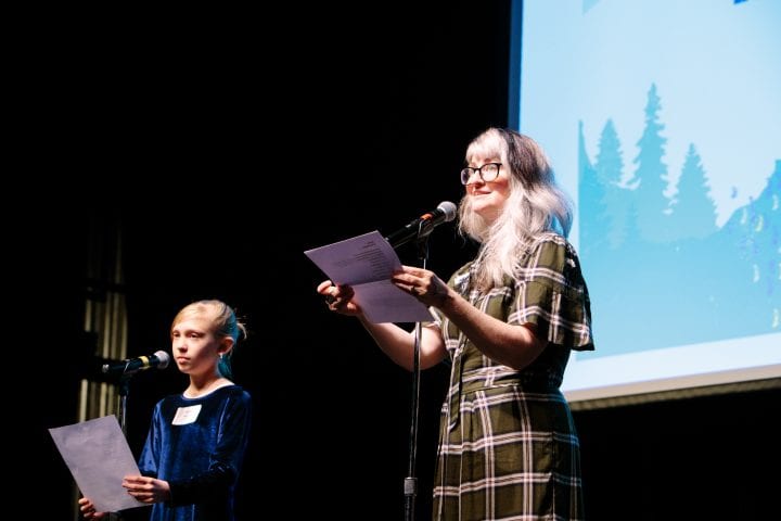 A woman stands at the microphone on stage, holding a piece of paper. A young girl stands at another microphone to the right.