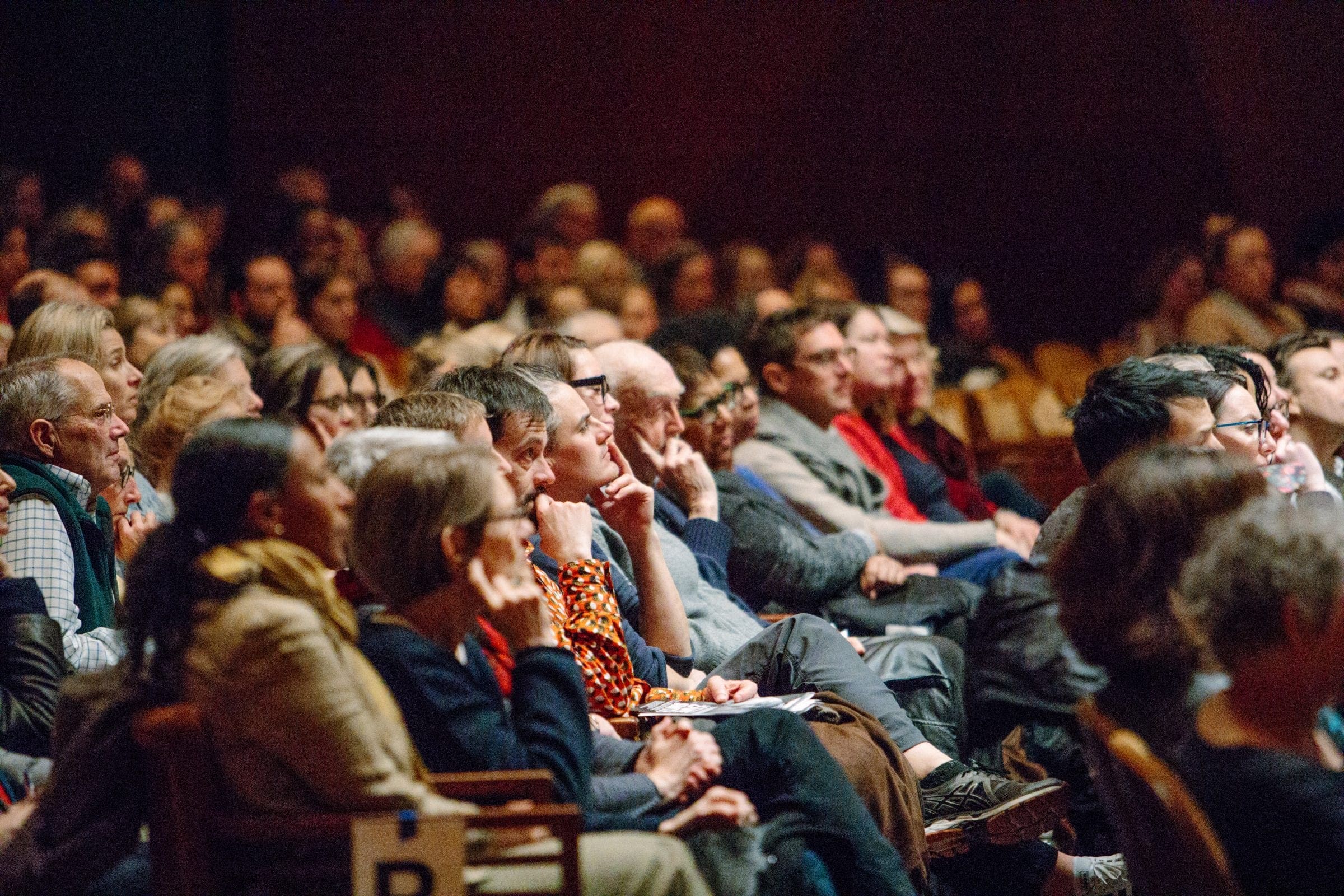 Several packed rows of an auditorium are pictured; several people have their elbows resting on their knees with their hands on their chins, their faces turned towards the stage.