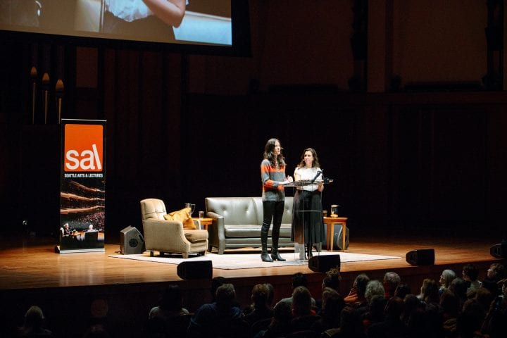 Two women with long, dark hair stand at a lucite lectern in front of a full Benaroya Hall, a brigh SAL banner and a living room stage set-up behind them.