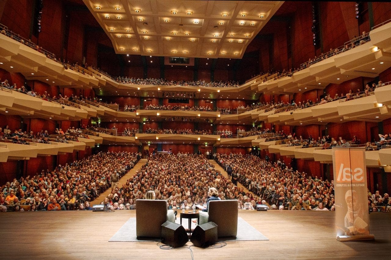 Two speakers sit in armchairs on a brightly-lit stage, with a fully-lit Benaroya Hall behind them; every seat looks full.