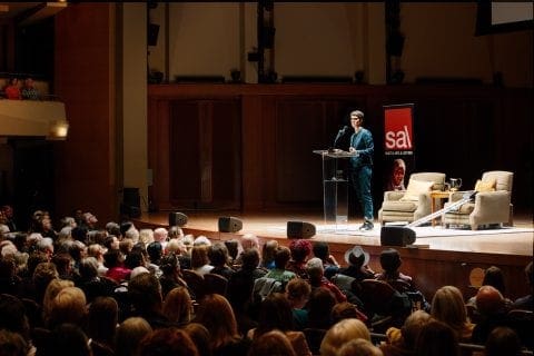 Rachel Maddow, in a blue blazer, stands at a lucite lectern, as the front rows of a crowd listen to her speak.