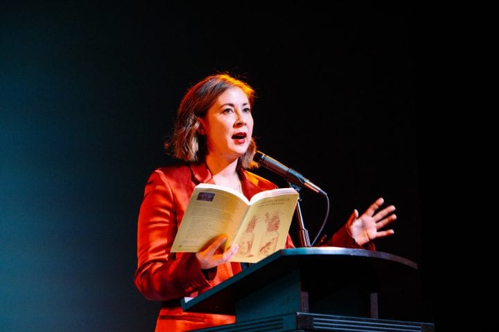 Paisley Rekdal, wearing a red satin suit, reads from her book at a lectern, one hand gesturing. Her gaze is cast upwards.