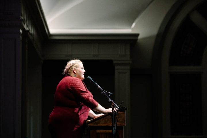 Lindy West, hair braided and wearing a red ribbed sweater, stands at a podium, with both hands powerfully placed upon it. Dramatic light shines down onto her as she speaks into a microphone.