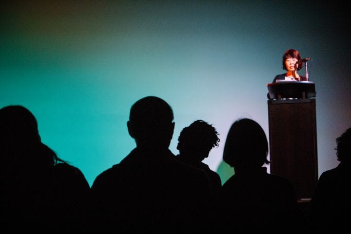 The shadowy foreground of the image shows the backlit heads of several audience members, listening to a short-haired woman who is reading and slightly out of focus in the background.