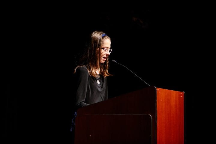 Lydia Ganz reads from a wood lectern, hair held back by a blue headband as she leans forward to look at her paper.