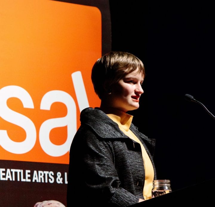 Lily Baumgart's profile is thrown into sharp relief against stage lights. Their hair is cropped short casts a shadow onto their cheek. They are wearing a yellow turtleneck with a wide-collared coat. A red SAL logo is behind the lectern where they're speaking.