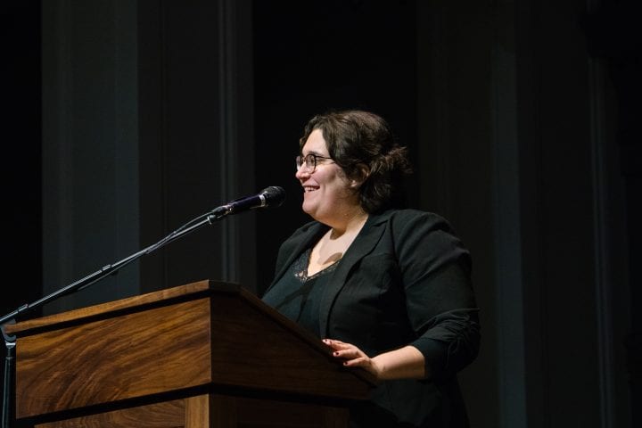 Carmen Maria Machado, wearing all black with short-cropped hair, stands a lectern and smiles during her lecture, gazing at a point off camera.