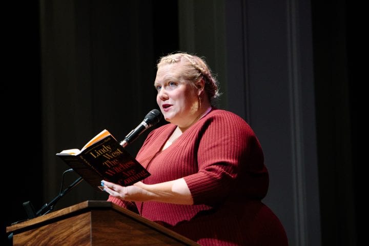Lindy West, wearing a red ribbed sweater, holds her book "The Witches are Coming" at a lectern, and reads from it while gazing up into the audience.