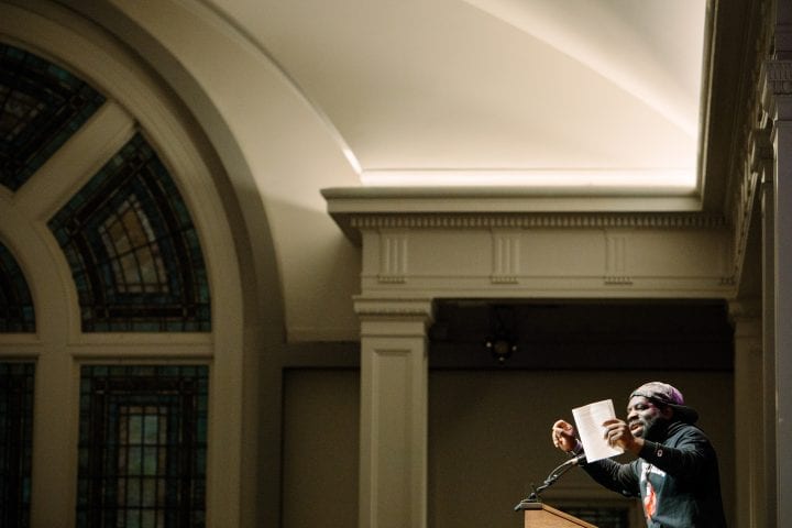 The interesting arched stained glass window and columns of Town Hall are the focus of this image, with Hanif Abdurraqib small in the right corner, emphatically gesturing at a lectern with a book in his hand. He is wearing a graphic, long-sleeved tee and a baseball cap.