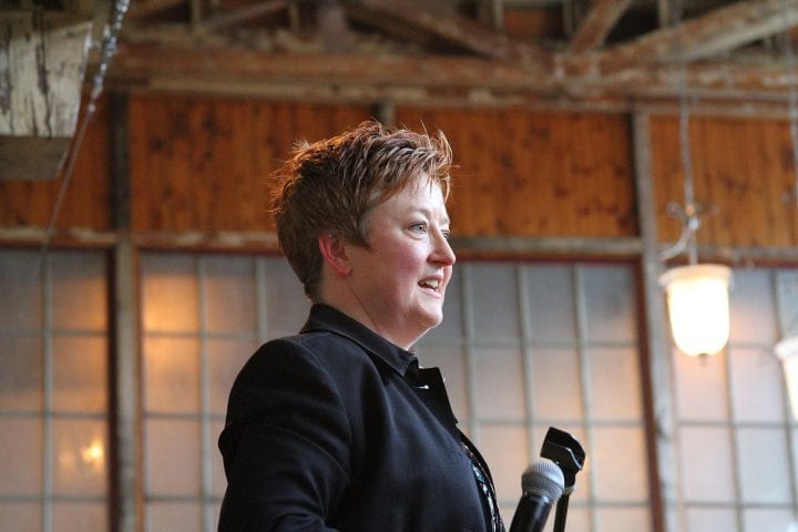 Amy Wheeler, with short-cropped, slightly spiky hair, stands in front of floor-to-ceiling, rustic-looking windows.