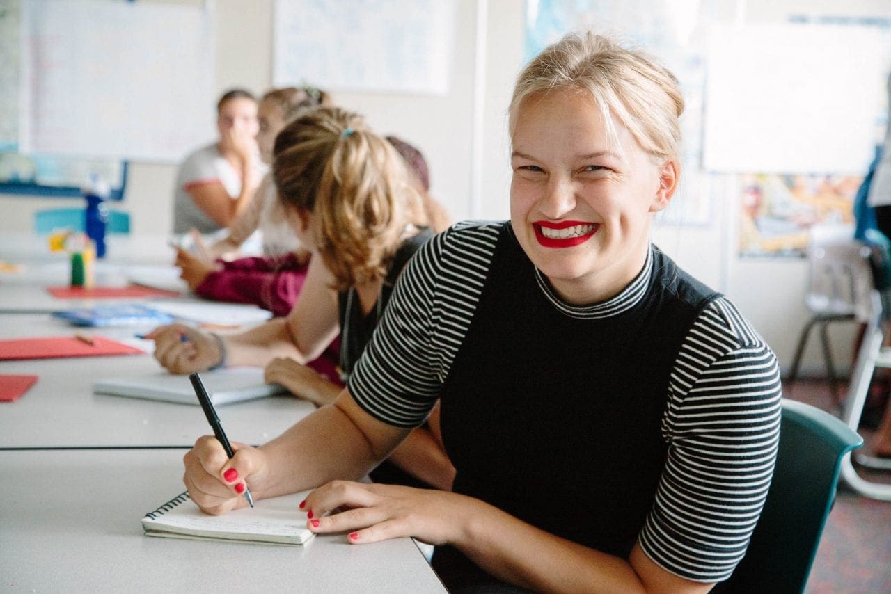 Lily Baumgaurt, at a WITS summer camp class, smiles while paused mid-writing, young writers in the background.