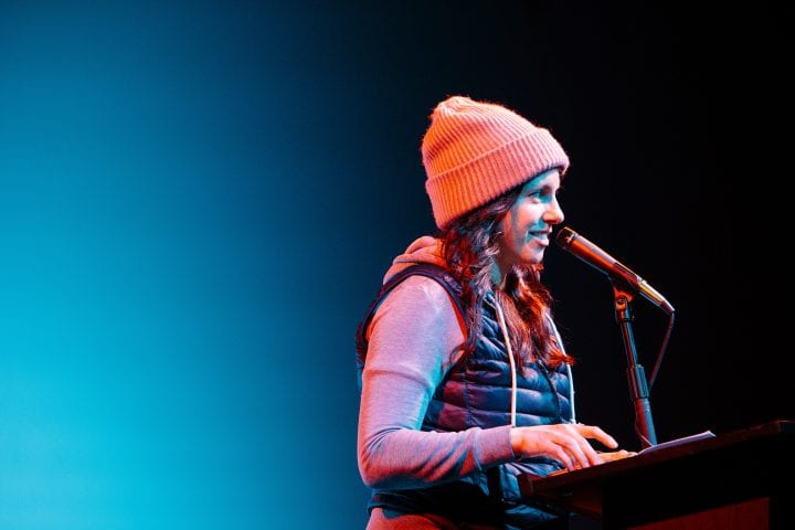 A woman stands onstage at the microphone, looking at the audience with a smile.