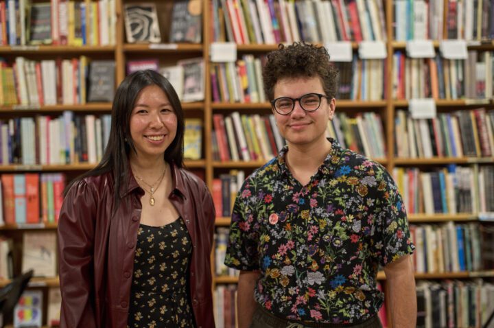 Two young adults smile in front of a full bookshelf.