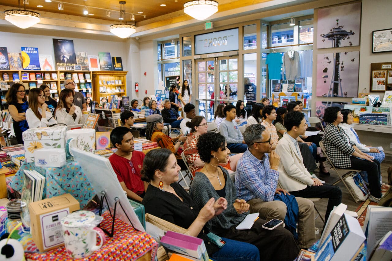 A group of 40 people seated and standing watch a presentation in a colorful, brightly lit bookstore.