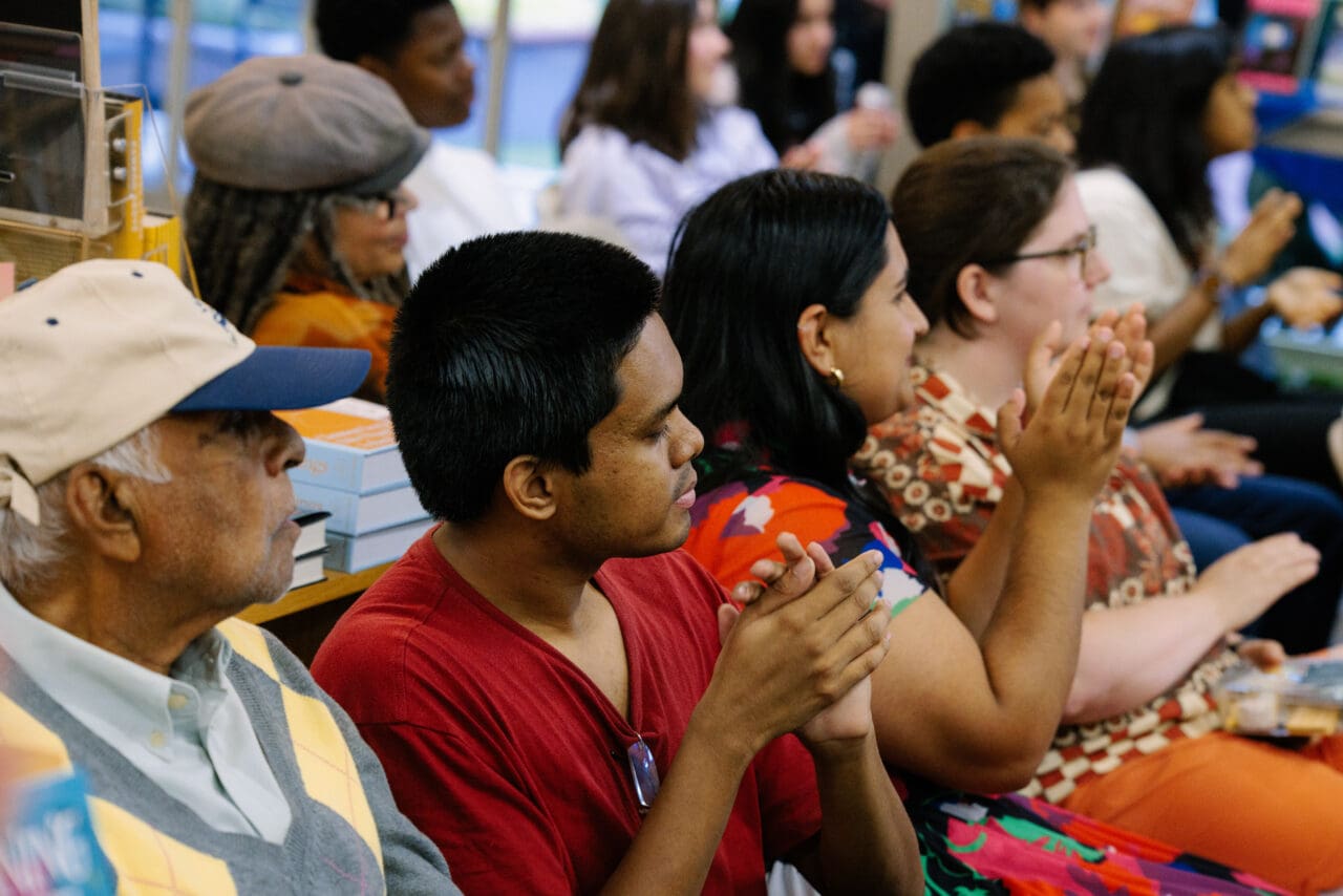 A row of supporters wearing bright clothes applaud while looking intently at a stage.