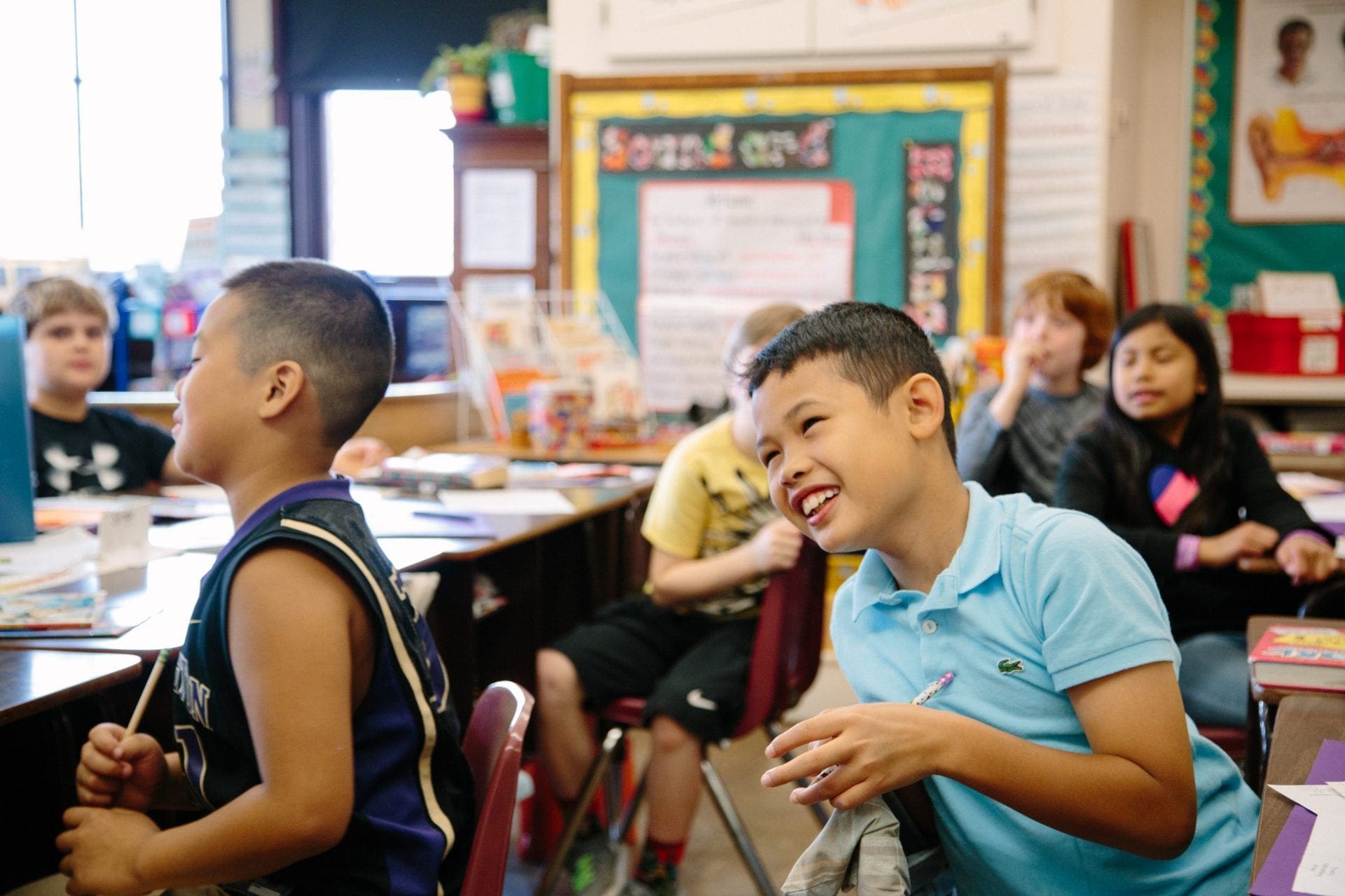 Elementary-aged students look to the front of a classroom. A boy, in the middle of the frame, leans forward in his chair towards what they're focused on, grinning.