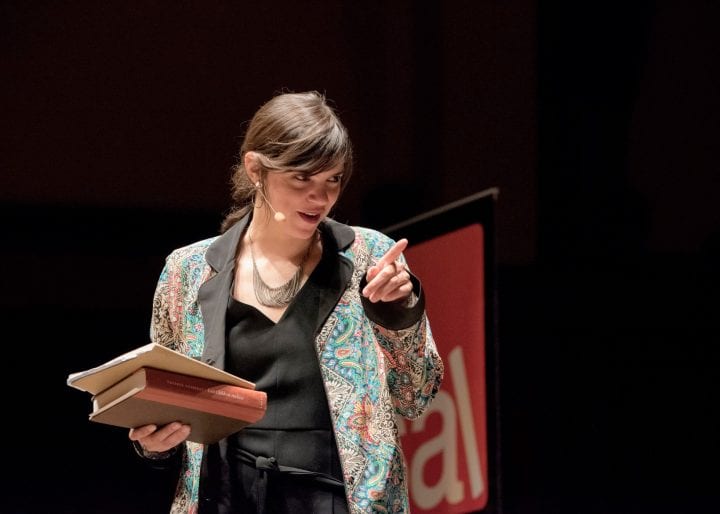 Valeria Luiselli, in a floral blazer, holds a stack of her books in her right hand, and points at someone off screen with her right.