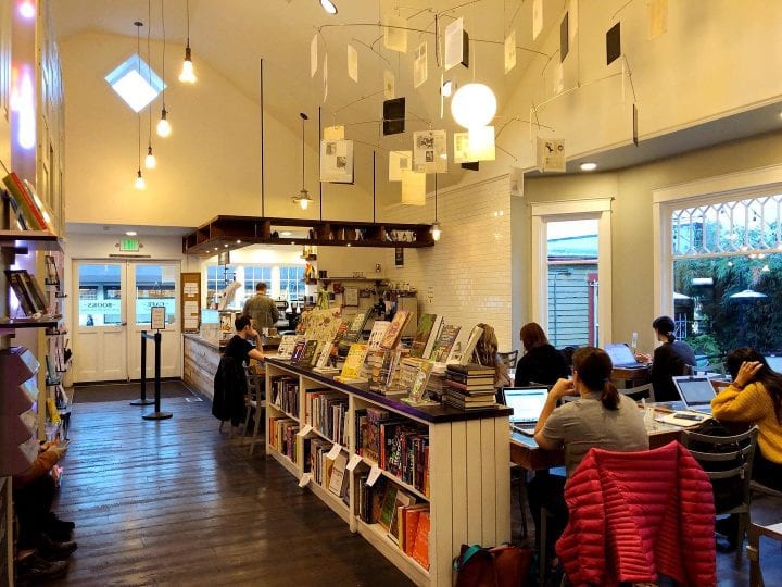 The warm, bright interior of Ada's Technical Books shows patrons sitting in the cafe area, on their computers behind a cookbook section, while a barista makes coffee in the background.