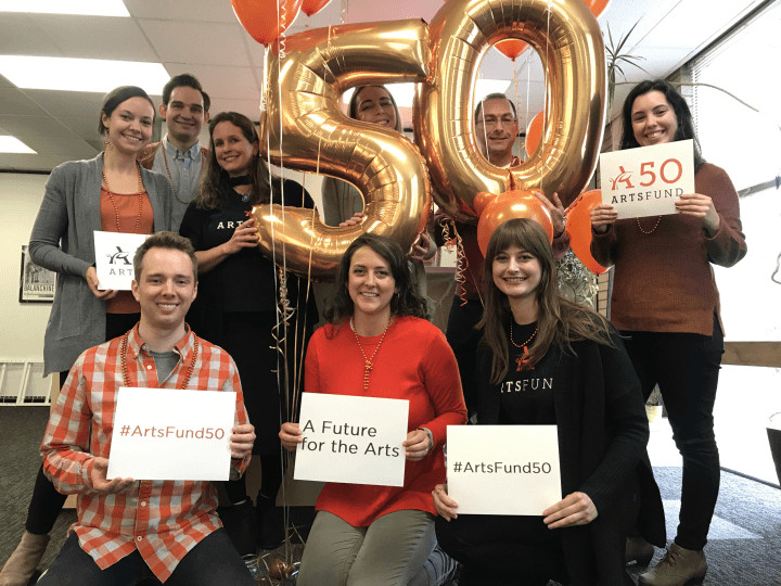 Nine ArtsFund staff member stand amid balloons, two of which read "50." The hold signs with the ArtsFund logos and hashtags.