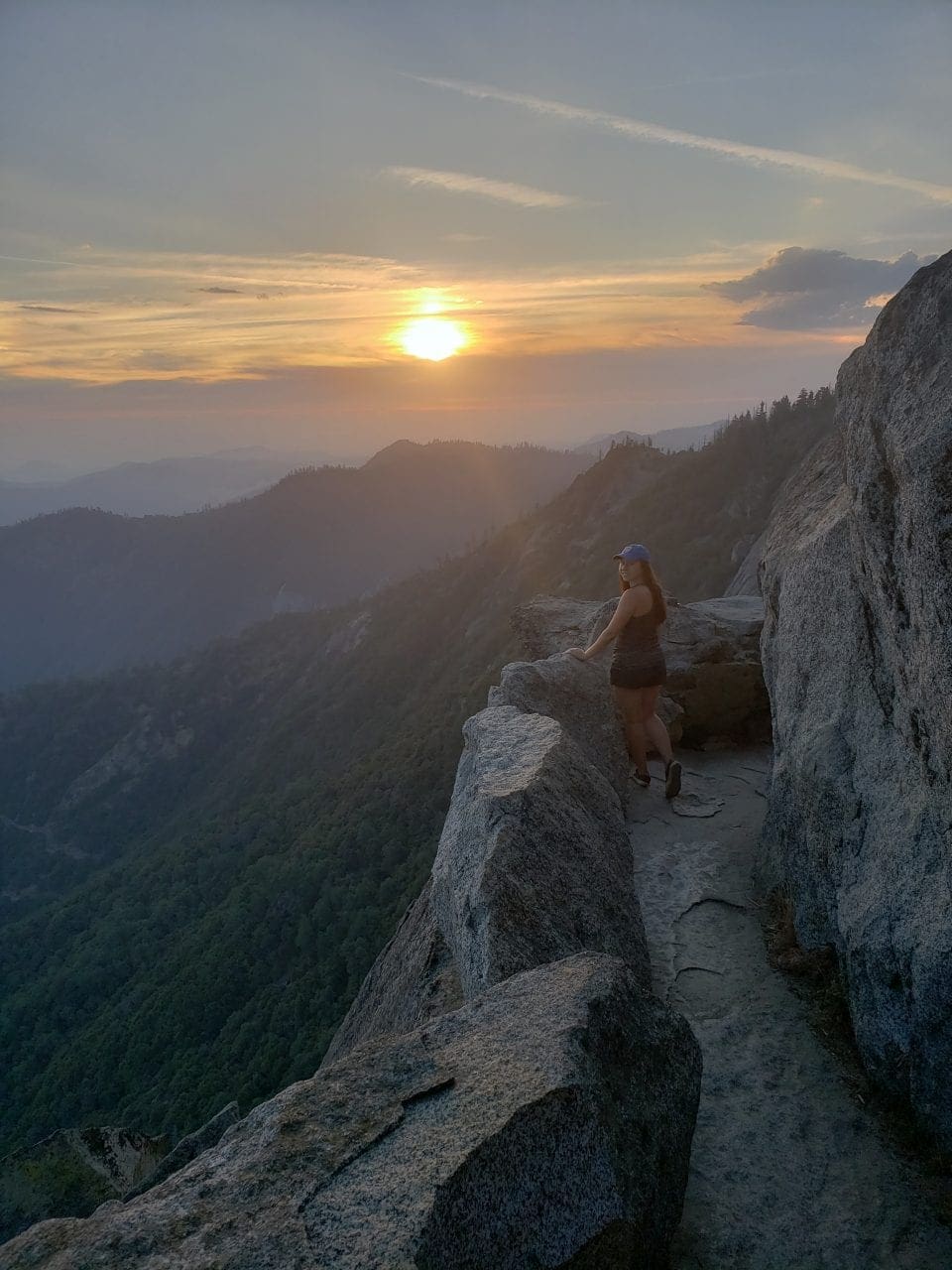 Bianca Glinskas stands on a cliff amid a sunrise, mountains in the background. She's leaning against a rock, small in the frame of the photo.