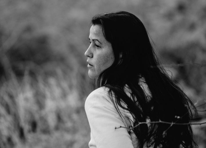A greyscale profile of Natalie Diaz, hair spilling over her shoulder, against a backdrop of long grasses