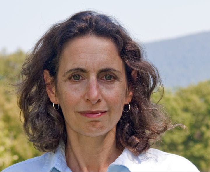 A close-up of Elizabeth Kolbert smiling, standing against a backdrop of unfocused trees.