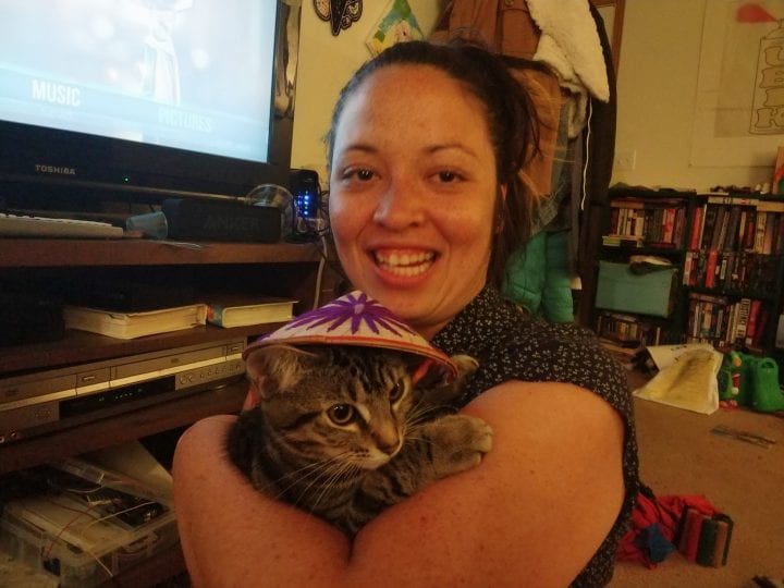 Natalie Howard is in her living room, holding her cat with one arm--her cat, a tabby, is wearing a miniature Vietnamese leaf hat.
