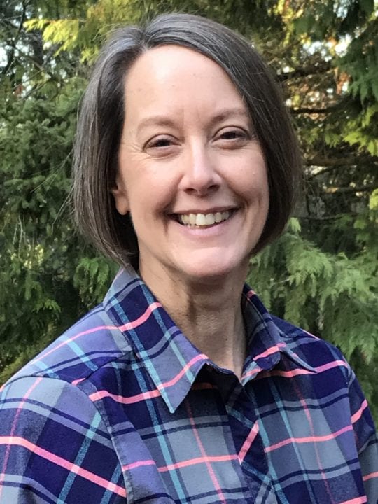 A close-up shot of Molly Suhr, smiling behind a backdrop of trees, wearing a purple plaid shirt.