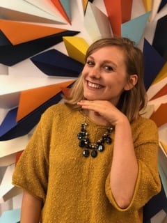 Cali Kopczick stands in front of a colorful geometric display, one hand propped under her face, which is smiling. She's wearing a mustard yellow sweater and a chunky necklace.