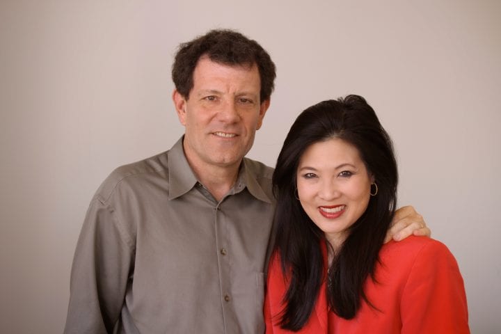 Nicholas Kristof has his arm over Sheryl WuDunn as they stand in front of a grey backdrop. Both are smiling.