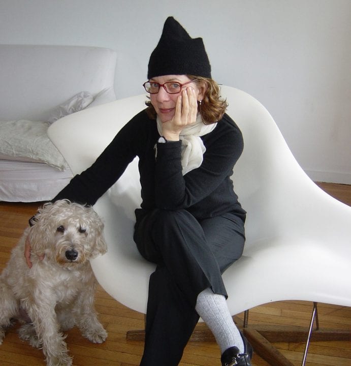 Maira Kalman, dressed in a pointy black hat, sits on a white modern chair, leaning on left elbow her right hand petting her curly-haired dog