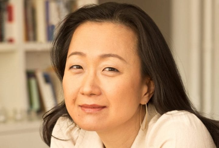 A close up of Min Jin Lee, wearing a white blouse, smiling and looking into the camera.