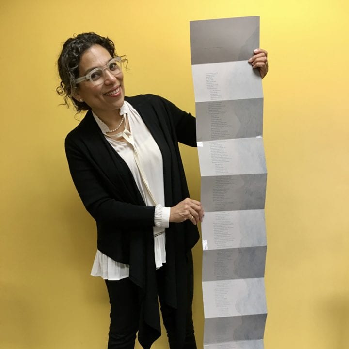 Claudia Castro Luna stands against a bright yellow backdrop, holding her series of poems, which unfolds accordian style, down from the top of her head all the way to the floor.