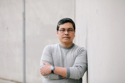 Rick Barot, dressed in a grey striped sweater, stand with arms crossed, leaning against a concrete wall