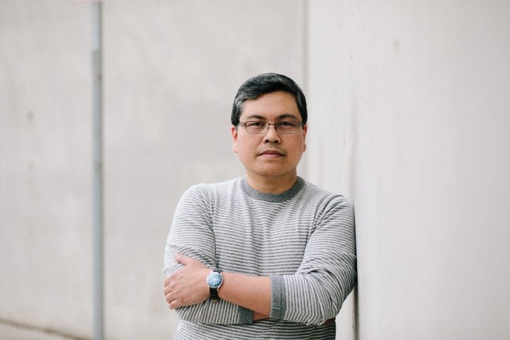 Rick Barot, dressed in a grey striped sweater, stand with arms crossed, leaning against a concrete wall