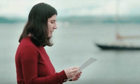Maia Pody stands in front of a blurred out background of a body of water, with boats behind her. She's looking down at a page she is reading aloud from.