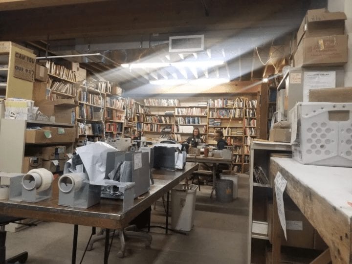A workspace at Books to Prisoners, with beams of light flooding down and touching shelves and boxes full of books. Two volunteers are working in the background.